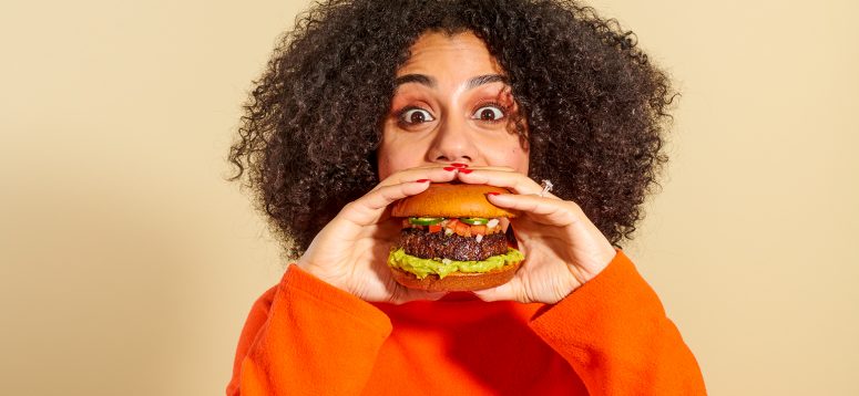 Female with dark curly hair, wearing an orange long sleeved shirt, holding a hamburger with tomatoes and jalapenos.